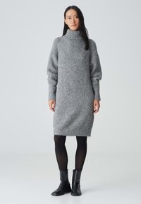 Graues Strickkleid mit hohem Rollkragen, langen Puffärmeln und geripptem Material. Kombiniert mit schwarzen Knöchelstiefeln und schwarzen Strumpfhosen.