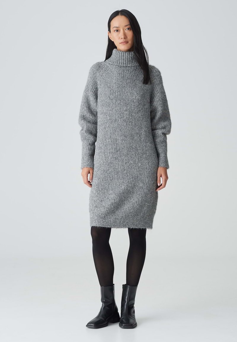 Graues Strickkleid mit hohem Rollkragen, langen Puffärmeln und geripptem Material. Kombiniert mit schwarzen Knöchelstiefeln und schwarzen Strumpfhosen.