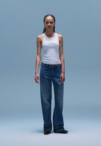 White sleeveless top paired with loose blue denim jeans and black shoes, set against a light blue background. Simple design and fit.