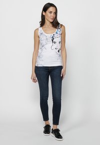 Top senza maniche bianco con stampa floreale nera e un volto. Abbinato a jeans skinny scuri e sneakers nere.