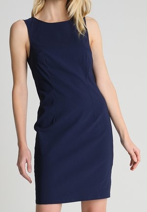 Mujer con un vestido azul marino ajustado sin mangas hasta la rodilla, de pie frente a un fondo liso gris claro.