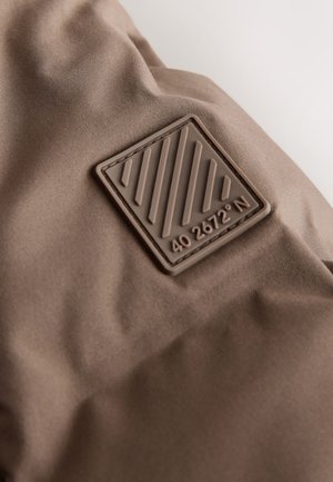 Tissu marron à la texture douce, avec une étiquette rectangulaire en caoutchouc marron ornée de lignes diagonales et du texte "40 2672° N."