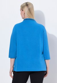 Top azul de manga 3/4 en una tela ligera y suave con un corte relajado y detalles de abertura lateral. Terminado con un diseño de cuello simple.