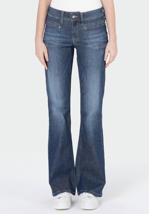 Femme portant un jean évasé en denim bleu taille mi-haute avec poches devant et baskets blanches, debout devant un fond uni.