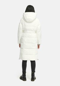 Witte pufferjas met een capuchon, ceintuurlijf en gewatteerde textuur. Gecombineerd met zwarte skinny jeans en enkellaarsjes. Achteraanzicht weergegeven.