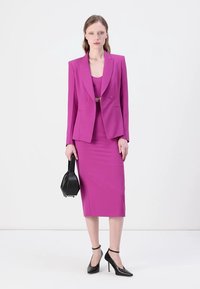 Blazer rose ajusté avec jupe crayon assortie, tissu lisse, revers nets et deux poches avant. Sac noir et talons complètent le look.
