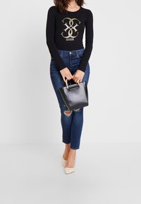Schwarzes Langarmoberteil mit goldenen Logo, blaue zerrissene Skinny-Jeans und eine kleine schwarze Handtasche mit goldenen Beschlägen, kombiniert mit beigen spitzen Absatzschuhen.