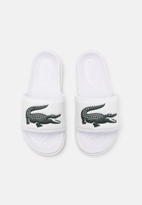 Tongs blanches en matériau synthétique, arborant un logo de crocodile noir sur la bride. Semelle intérieure texturée pour une meilleure adhérence et un confort accru.