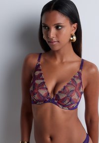 Bralette en dentelle violet avec des motifs floraux complexes en rouge et or. Il présente des bretelles ajustables et un détail décoratif de bouton au centre.