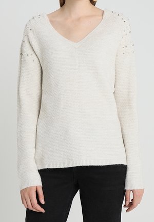 Femme portant un pull en maille crème avec un col en V et de petites perles semblables à des perles sur les épaules, associé à un pantalon noir.