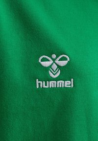 Logo hummel blanc brodé avec un bourdon stylisé sur un fond en tissu vert.