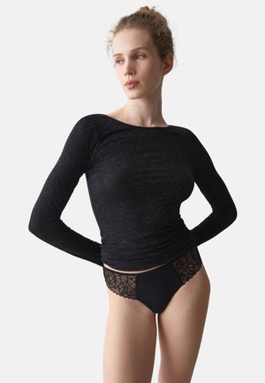 Kvinde med lys hud og hår sat op i knold bærer en tætsiddende sort bluse med lange ærmer og sort undertøj med blonder, kigger til siden med hænderne bag ryggen.