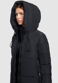 Sort dunjakke med stor hættekrave, lynlås foran og snoredetaljer. Tekstureret quiltet design med sidelommer.