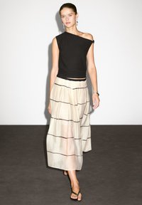 Haut asymétrique noir à manches courtes ; jupe crème fluide avec des rayures horizontales noires ; portée avec des tongs noires simples.