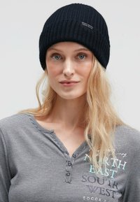 Black ribbed knit beanie with a logo tag. Model wears a gray striped long-sleeve shirt featuring text and button details.