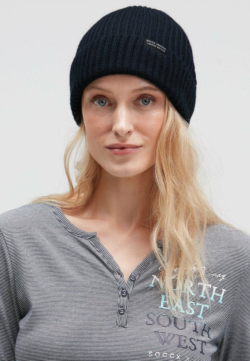 Black ribbed knit beanie with a logo tag. Model wears a gray striped long-sleeve shirt featuring text and button details.