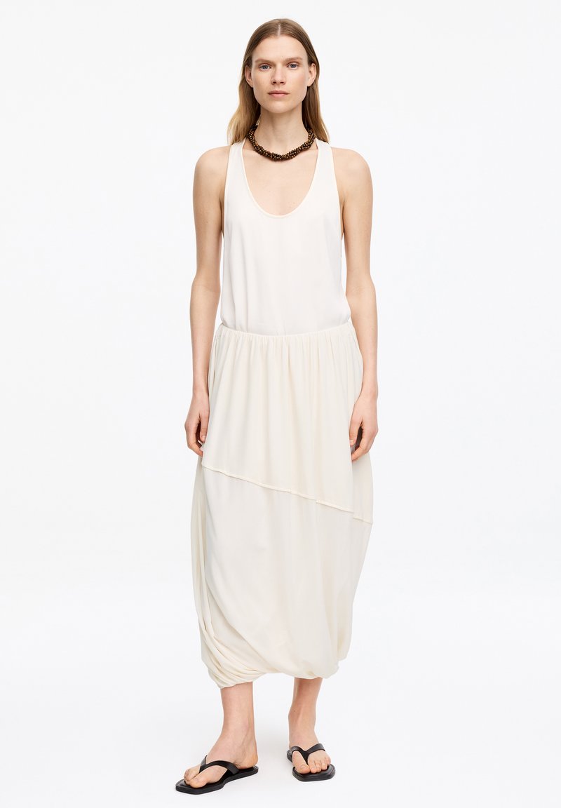 Femme portant une robe blanche sans manches avec une jupe à volants et des tongs noires, accessoirisée d'un gros collier de perles marron, debout sur fond blanc.