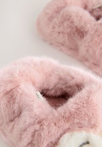Chaussons en peluche roses avec une texture douce et duveteuse et une forme arrondie. Présentent un visage d'animal décoratif à l'avant et une étiquette de marque à l'intérieur.