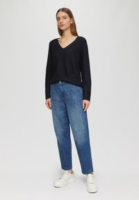 Schwarzer V-Ausschnitt-Pullover, lockere Passform, kombiniert mit hellblauen High-Waist-Jeans. Weiße Plateausneaker runden das Outfit ab, vor einem neutralen Hintergrund.