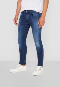 Jeans ajustados de color azul oscuro con textura desgastada, diseño clásico de cinco bolsillos, herrajes plateados y detalles de costura blanca. Combinados con zapatillas blancas.
