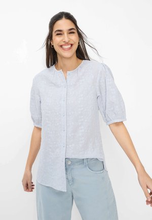 Junge Frau lächelnd, trägt eine hellblaue bestickte Bluse mit Knöpfen und Puffärmeln, kombiniert mit hellblauen Hosen.