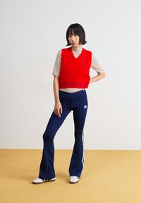 Piros kötött mellény bézs garbó felett, navy kicsípett nadrág fehér csíkokkal, és fehér sportcipő fekete részletekkel.