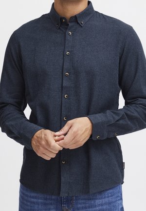 Homme portant une chemise bleu foncé à manches longues boutonnée avec col boutonné, ajustant les poignets, montré du cou aux hanches.