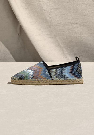 Chaussure espadrille avec motif zigzag multicolore, bordure noire, semelle en jute tressé, présentée sur une surface claire avec un fond en tissu beige.