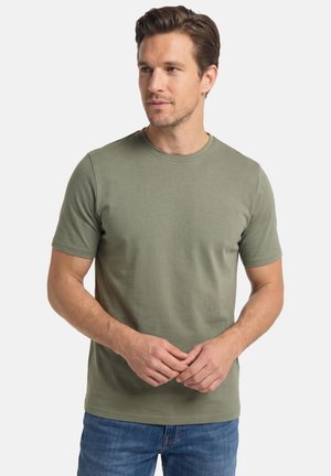 Mand med brunt hår iført en olivengrøn t-shirt med korte ærmer og blå jeans, der kigger let til siden med foldede hænder.