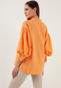 Oranžs puff-sleeve blūze vieglā audumā. Krekliņa apkakle ar pogām, ikdienas griezums un noapaļots apakšmalas dizains. Saskaņojot ar nūda legingiem.