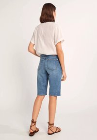 Shorts en denim bleu style bermuda avec une coupe droite, cinq poches et un ourlet retroussé. Associés à un chemisier blanc ample et des sandales marron.