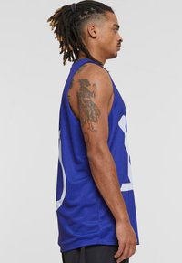 Canotta da basket blu in mesh con design del logo bianco, senza maniche, taglio ampio e superficie testurizzata, indossata da una persona con tatuaggi.