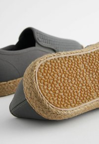 Harmaat kangas liukuvat kengät, joissa on kudotut jute-korostukset pohjassa. Teksturoitu kumipohja, jossa on pistekuvio, tarjoaa pitoa.