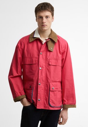 Noor mees, kes kannab punast nööbitavat jopet, millel on pruun krae ja mansetid ning eest nööbitavad taskud, seisab ühevärvilise valge tausta ees.