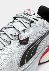 Γκρι αθλητικό παπούτσι με μαύρες λεπτομέρειες, υφή από δίχτυ, και κόκκινες διακοσμήσεις. Διαθέτει επίπεδους κορδόνια και επενδεδυμένο κολάρο για στήριξη.