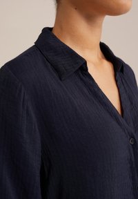 Jūras spēku zila pogām apģērba krekls, izgatavots no teksturēta auduma. Iekļauj salocītu apkakli un priekšējo pogu aizdari, uzsverot brīvo stilu.