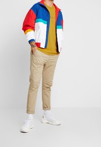 Hombre con chaqueta multicolor azul, roja y blanca sobre una camiseta mostaza, pantalones beiges remangados, calcetines blancos y zapatillas blancas, con una mano en el bolsillo.
