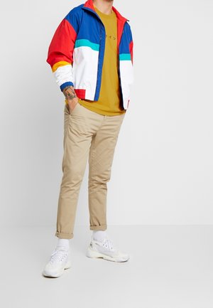 Hombre con chaqueta multicolor azul, roja y blanca sobre una camiseta mostaza, pantalones beiges remangados, calcetines blancos y zapatillas blancas, con una mano en el bolsillo.