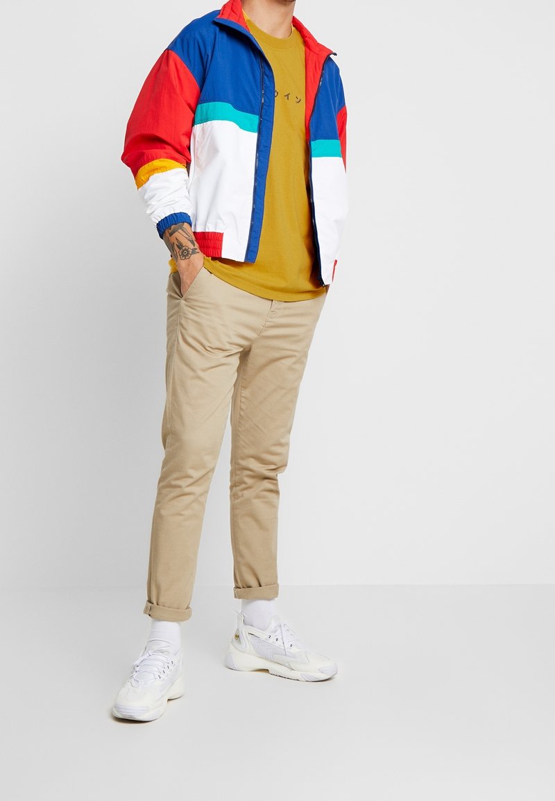 Hombre con chaqueta multicolor azul, roja y blanca sobre una camiseta mostaza, pantalones beiges remangados, calcetines blancos y zapatillas blancas, con una mano en el bolsillo.