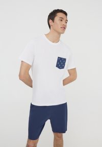 T-shirt bianco a maniche corte con taschino blu navy decorato con motivi di palme bianche, abbinato a pantaloni corti blu navy. Design semplice e casual.