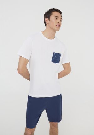 T-shirt bianco a maniche corte con taschino blu navy decorato con motivi di palme bianche, abbinato a pantaloni corti blu navy. Design semplice e casual.