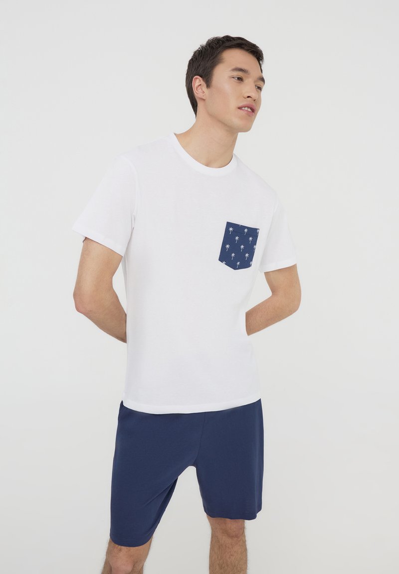 T-shirt bianco a maniche corte con taschino blu navy decorato con motivi di palme bianche, abbinato a pantaloni corti blu navy. Design semplice e casual.