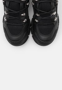 Svarta sneakers med en texturerad meshöverdel, prydda med silverfärgade öglor, tjocka svarta sulor och svarta snören. Inga framträdande varumärken synliga.