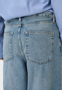 Gaiši zili džinsi ar augstu jostasvietu, divi aizmugurējie kabatas un klasiskā taisnā kāju dizaina, attēlā no sānu skatpunkta.