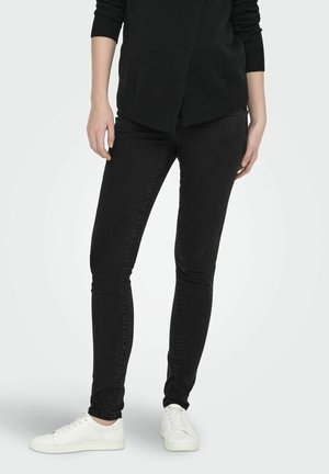 Personne portant un jean skinny noir, un haut à manches longues noir et des baskets blanches, debout devant un fond clair uni.