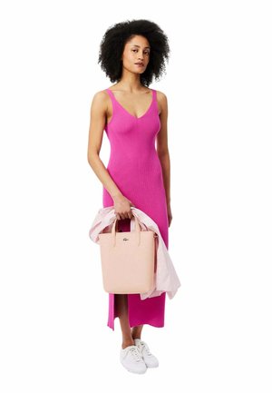 Robe tricotée côtelée fuchsia avec un décolleté en V, longueur au genou. Le modèle porte un sac fourre-tout rose clair et tient une chemise rayée. Baskets blanches.