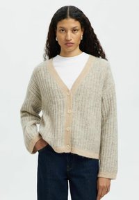 Värikäs beige ribbikardigan V-kaula-aukolla, jossa on vaaleanbeige kontrastisivusto ja viisi nappia, yhdistetty valkoiseen paitaan ja tummiin farkkuihin.