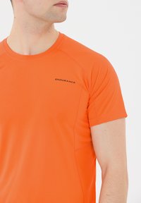 Oranssi urheilupaita lyhyillä hihoilla, valmistettu teksturoidusta kankaasta. Ominaisuuksiin kuuluu pyöreä pääntie ja "ENDURANCE"-logo rinnassa.