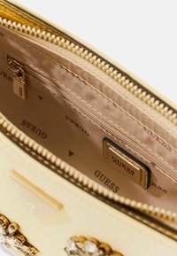 Beżowe wnętrze torebki Guess z podszewką z materiału z logo, złotym suwakiem i dekoracyjnymi kryształowymi ozdobami na zewnątrz.