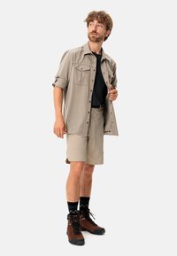 Camicia da esterno beige con maniche arrotolate, t-shirt nera sotto, pantaloni corti beige e scarponi da trekking marroni con calze nere a fantasia.